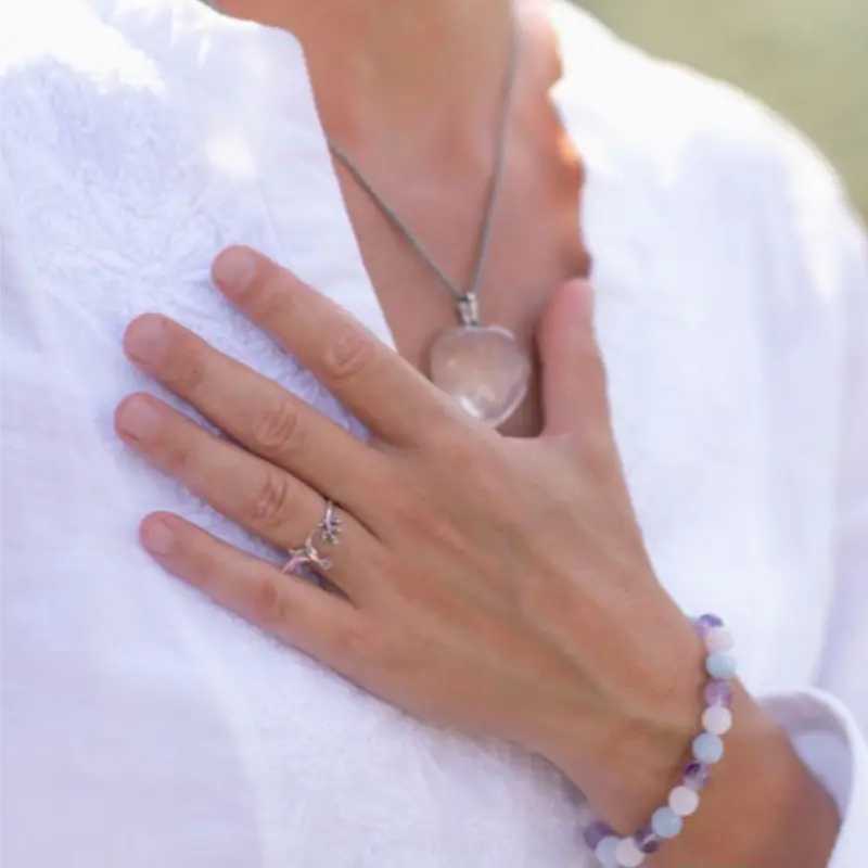 HandaufsHerz Frau in weisser Bluse legt ihre Hand auf die Brust, am Hals eine Kette mit einem herzförmigen Kristall als Anhänger.