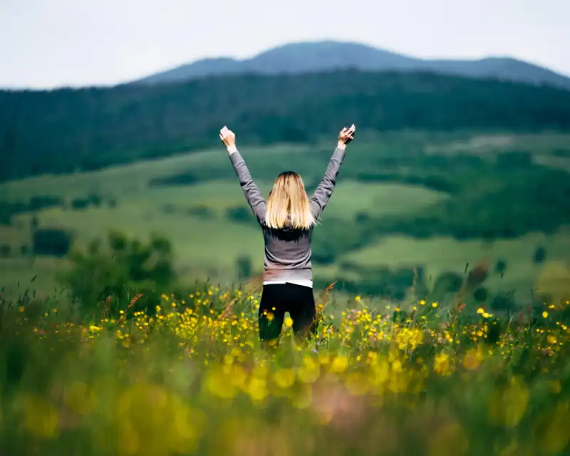 PE_success Junge Frau mit blonden Haaren auf einer Blumenwiese mit Hügeln im Hintergrund reckt die Arme als Zeichen des Erfolgs in die Höhe. Von hinten betrachtet
