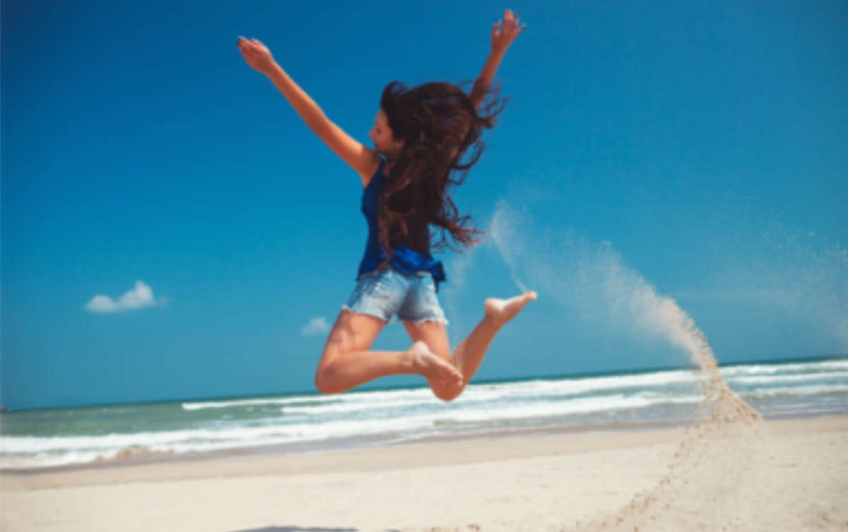 Eine junge Frau in Jeans Shorts springt mit ausgebreteiteten Armen an einem Sandstrand in die Luft und freut sich des Lebens.